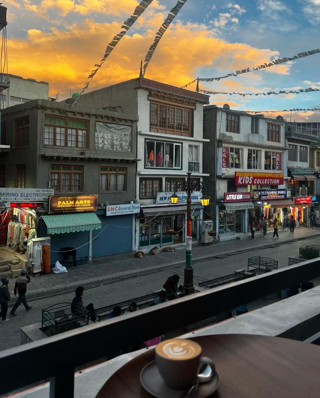 Prayer flags strung over a mountain bazaar at sunset, a cup of coffee on the table in the foreground